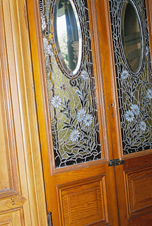 [Two wooden doors with the upper two thirds made of stained glass. There is an oval clear glass section in each of the two panes. The rest of the windows are a daisy motif with white, yellow, red, and green glass.]